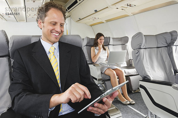 Deutschland  Bayern  München  Geschäftsleute  die an iPad und Laptop in der Business-Class-Flugzeugkabine arbeiten  lächelnd