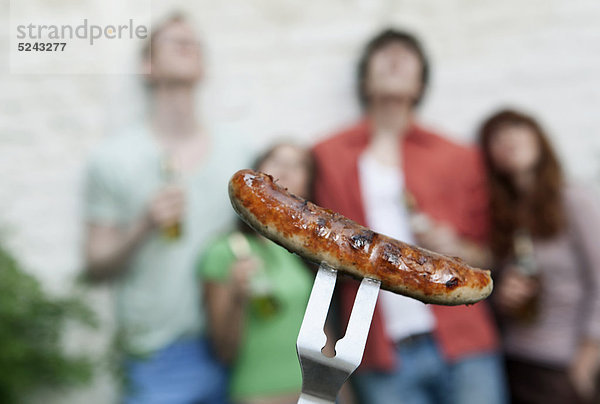 Deutschland  Berlin  Nahaufnahme der Grillwurst auf der Gabel mit Freunden im Hintergrund