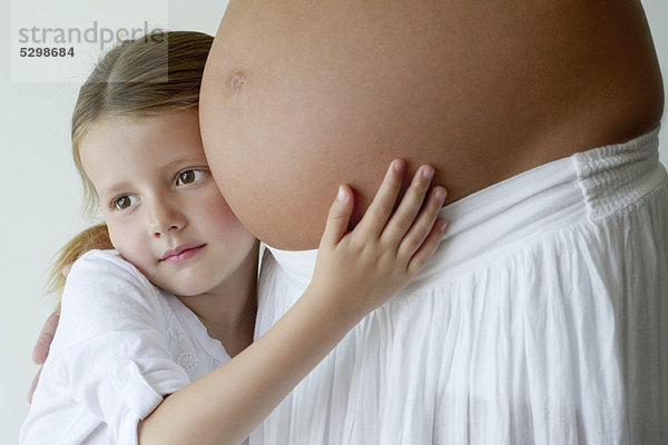 Mädchen  das den schwangeren Bauch der Mutter umarmt.