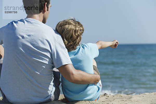 Vater und Sohn sitzen zusammen am Strand  Junge zeigt auf das Meer.