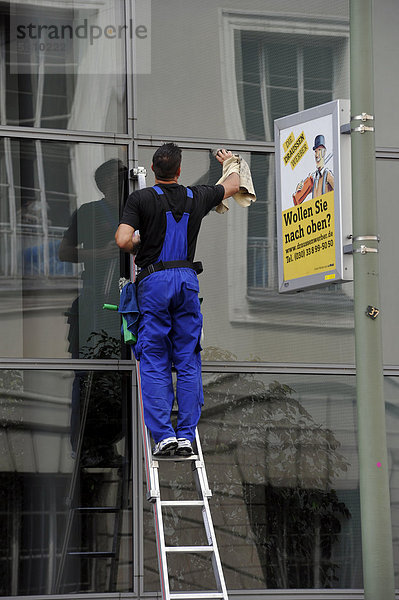 Glasfassadenreiniger auf Leiter bei der Arbeit  neben Werbebanner Willst Du hoch hinaus?  Berlin  Deutschland  Europa  ÖffentlicherGrund