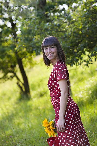 Junge Frau  25 Jahre  mit getupftem Kleid und Sonnenblume  steht in Wiese