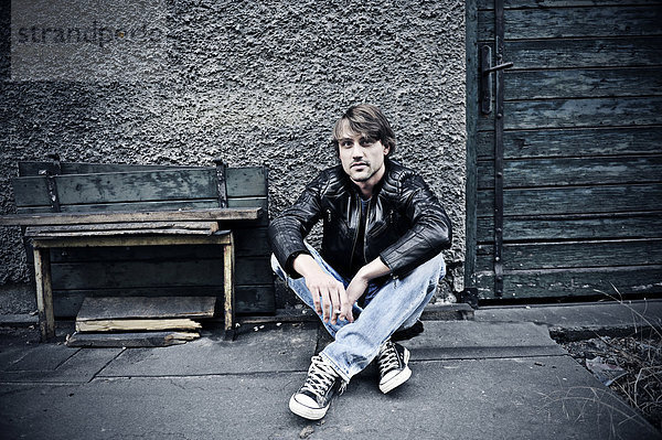Mann in Jeans und Lederjacke mit Zigarette sitzt vor alter Mauer