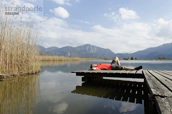 Mann liegt auf Steg am Eichsee im Kochler Moor bei Schlehdorf Kochel  mit dem Herzogstand und Jochberg  Oberbayern  Bayern  Deutschland  Europa