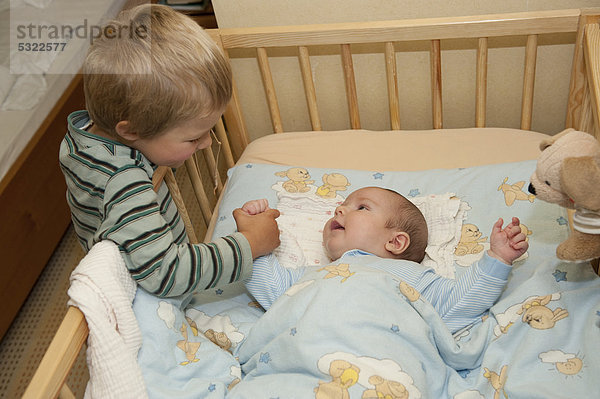 Junge  3 Jahre  spielt mit seinem kleinen Bruder  4 Monate  am Gitterbettchen  hält seine Hand