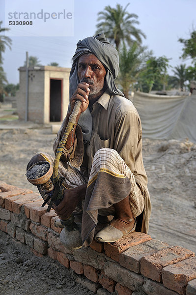 Mann sitzt mit großer Pfeife auf einer Ziegelmauer  Dorf Basti Lehar Walla  Punjab  Pakistan  Asien