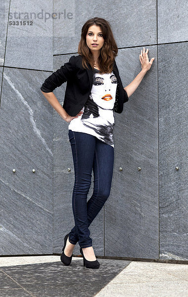 Junge Frau in Top mit Porträtmotiv und Jeans posiert an grauer Wand