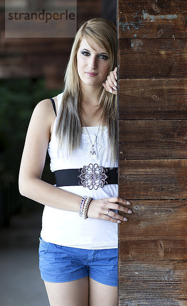 Junge Frau mit langen blonden Haaren posiert stehend an Holzwand