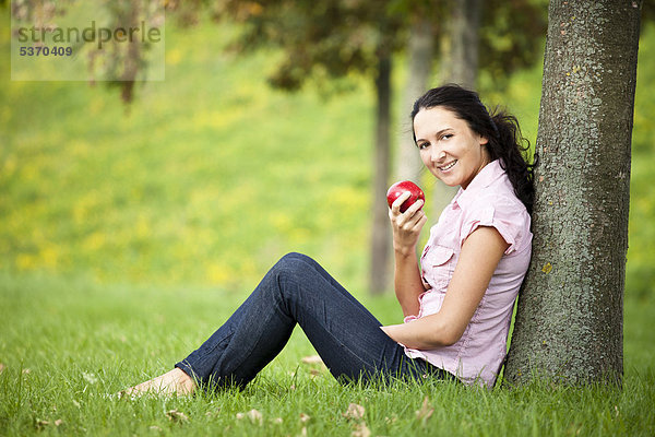 Junge Frau lehnt an einem Baum  ißt einen roten Apfel