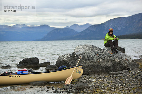 Junge Frau sitzt auf einem Felsen  Kanu und Holzpaddel am Ufer  Kusawa Lake  die Berge dahinter  Yukon Territory  Kanada