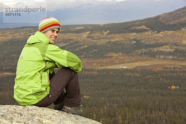 Junge Frau sitzt auf einem Felsen auf einem Berg  rastet  genießt Aussicht  subalpine Tundra  Indian Summer  Herbst  in der Nähe des Fish Lake  Yukon Territory  Kanada