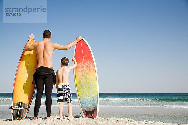 Vater und Sohn mit Surfbrettern  die aufs Meer hinausblicken.