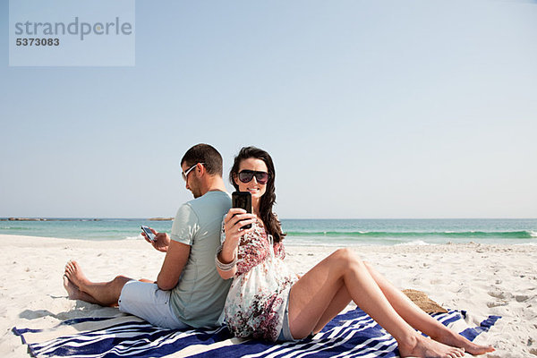 Paar sitzt Rücken an Rücken am Strand und benutzt dabei Mobiltelefone.