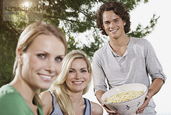 Italien  Toskana  Magliano  Junger Mann hält Schale mit Pasta und Frauen im Vordergrund  lächelnd  Portrait