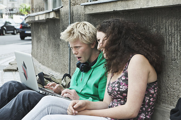 Deutschland  Berlin  Teenagerpaar auf dem Bürgersteig sitzend mit Laptop