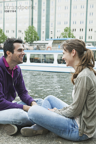 Deutschland  Berlin  Paar sitzend von Angesicht zu Angesicht am Flussufer