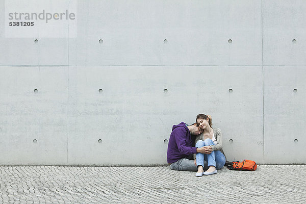 Deutschland  Berlin  Paar sitzend vor großer Mauer auf Bürgersteig