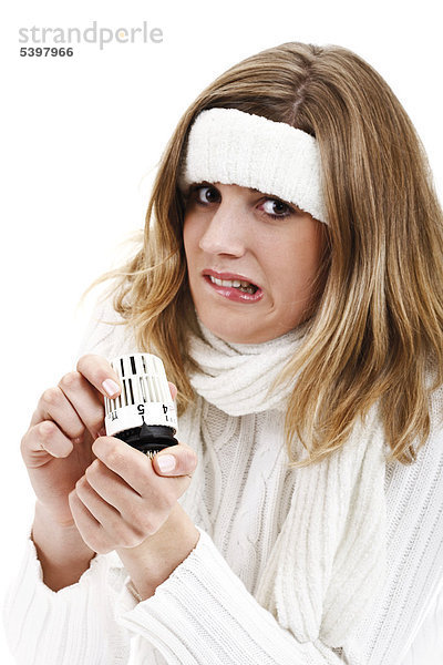 Junge Frau im weißen Rollkragenpullover mit Stirnband hält frierend Heizungsthermostat