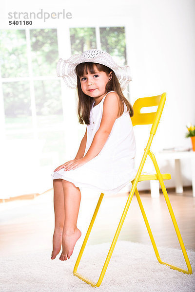 Kleines Mädchen mit weißem Kleid und Hut  auf Stuhl sitzend