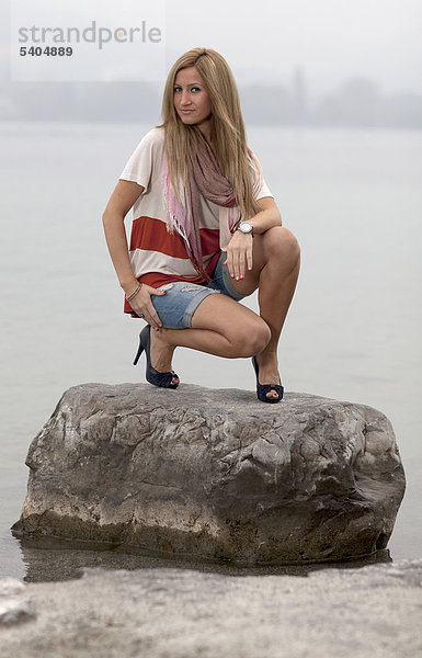 Junge blonde Frau in Hotpants posiert auf Stein im Wasser