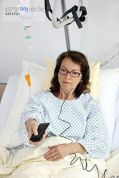Patientin im Krankenbett  bedient mit der Fernbedienung einen Fernseher  hört den Ton über Ohrhörer  Krankenhaus