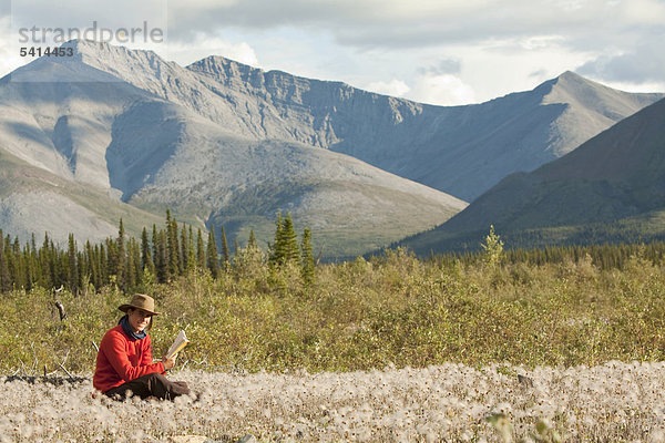Junge Frau liest ein Buch  entspannt  sitzt im Wollgras  Peel Watershed  hinten die nördlichen Mackenzie Mountains  Yukon Territory  Kanada