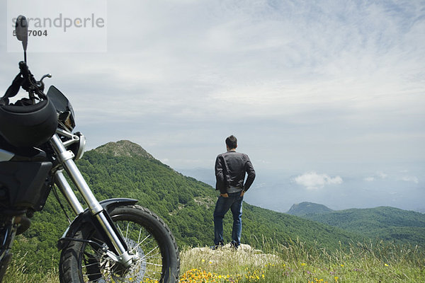 Mann auf Felsen stehend mit Blick auf die Berge  Motorrad im Vordergrund  Rückansicht