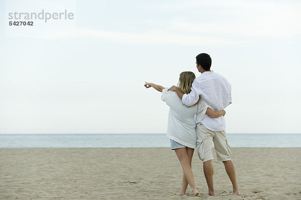 Ein Paar  das zusammen am Strand spazieren geht  eine Frau  die auf das Meer zeigt.