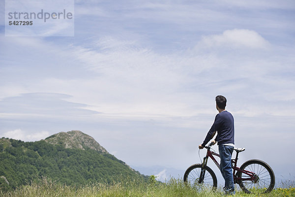 Mann steht am Mountainbike  genießt die Aussicht auf die Berge  Rückansicht