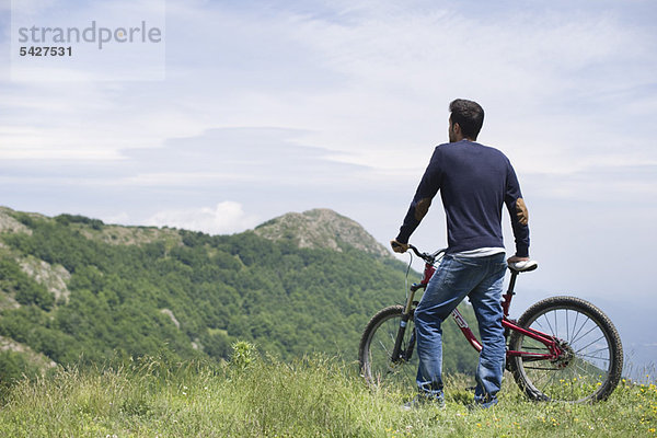 Mann steht am Mountainbike  genießt die Aussicht auf die Berge  Rückansicht