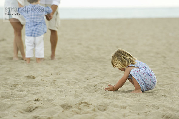 Kleines Mädchen spielt im Sand am Strand  Familie im Hintergrund