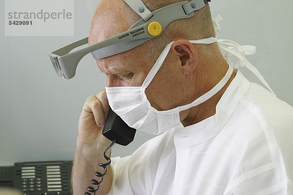 Zahnarzt mit Mundschutz und Lupenbrille am Telefon