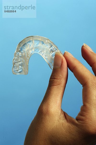 Bruxismus - bruxism - das meist unbewusste nächtliche Knirschen mit den Zähnen - durch das Knirschen mit den Zähnen treten in den meisten Fällen Fehlbelastungen der Zähne bzw. Zahngruppen oder Kiefergelenksbeschwerden auf - um eine Entlastung der Zähne und eine Gelenkschonung bei Kieferarthropathie zu erreichen wird eine individuell angepasste Kunststoffplatte   Knirscherschiene   Aufbissbehelf ( Okklusionsschiene oder Relaxierungsschiene ) für den Patienten angefertigt - diese sollte vorwiegend nachts getragen werden - Ursachen können psychischer ( Stress )   physischer ( Skeletterkrankungen ) oder neurologischer ( MS Multiple Sklerose ) Natur sein - eine Frau hält eine Knirscherschiene für den Oberkiefer zwischen den Fingern