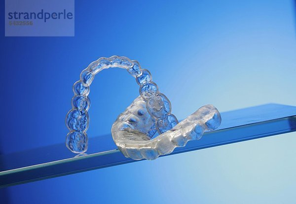 Beißschienen für Oberkiefer und Unterkiefer auf Glasplatten vor blauem Hintergrund ( Orthoarch - lat.) - Bruxismus - bruxism - das meist unbewusste nächtliche Knirschen mit den Zähnen - durch das Knirschen mit den Zähnen treten in den meisten Fällen Fehlbelastungen der Zähne bzw. Zahngruppen oder Kiefergelenksbeschwerden auf - um eine Entlastung der Zähne und eine Gelenkschonung bei Kieferarthropathie zu erreichen wird eine individuell angepasste Kunststoffplatte   Knirscherschiene   Aufbissbehelf ( Okklusionsschiene oder Relaxierungsschiene ) für den Patienten angefertigt - diese sollte vorwiegend nachts getragen werden - Ursachen können psychischer ( Stress )   physischer ( Skeletterkrankungen ) oder neurologischer ( MS Multiple Sklerose ) Natur sein