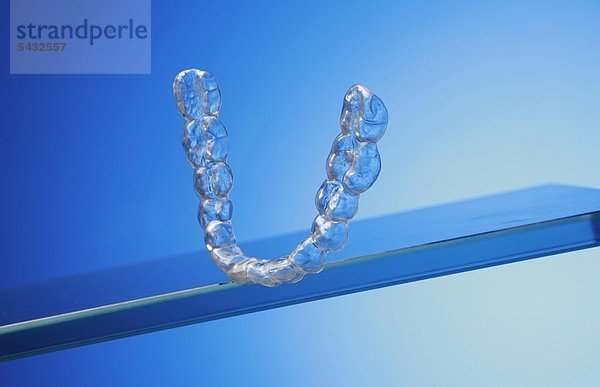 Untere Beißschiene für Unterkiefer auf Glasplatte vor blauem Hintergrund ( Orthoarch - lat.) - Bruxismus - bruxism - das meist unbewusste nächtliche Knirschen mit den Zähnen - durch das Knirschen mit den Zähnen treten in den meisten Fällen Fehlbelastungen der Zähne bzw. Zahngruppen oder Kiefergelenksbeschwerden auf - um eine Entlastung der Zähne und eine Gelenkschonung bei Kieferarthropathie zu erreichen wird eine individuell angepasste Kunststoffplatte   Knirscherschiene   Aufbissbehelf ( Okklusionsschiene oder Relaxierungsschiene ) für den Patienten angefertigt - diese sollte vorwiegend nachts getragen werden - Ursachen können psychischer ( Stress )   physischer ( Skeletterkrankungen ) oder neurologischer ( MS Multiple Sklerose ) Natur sein