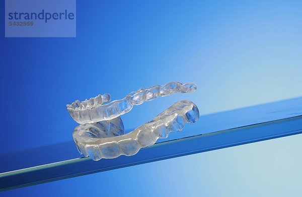 Beißschienen für Oberkiefer und Unterkiefer auf Glasplatte vor blauem Hintergrund ( Orthoarch - lat.) - Bruxismus - bruxism - das meist unbewusste nächtliche Knirschen mit den Zähnen - durch das Knirschen mit den Zähnen treten in den meisten Fällen Fehlbelastungen der Zähne bzw. Zahngruppen oder Kiefergelenksbeschwerden auf - um eine Entlastung der Zähne und eine Gelenkschonung bei Kieferarthropathie zu erreichen wird eine individuell angepasste Kunststoffplatte   Knirscherschiene   Aufbissbehelf ( Okklusionsschiene oder Relaxierungsschiene ) für den Patienten angefertigt - diese sollte vorwiegend nachts getragen werden - Ursachen können psychischer ( Stress )   physischer ( Skeletterkrankungen ) oder neurologischer ( MS Multiple Sklerose ) Natur sein