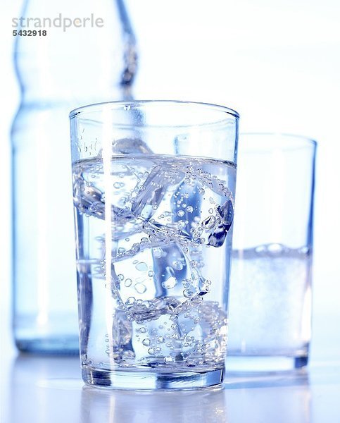 Glas gefüllt mit Wasser und Eiswürfeln - dahinter ein weiteres Glas gefüllt mit Wasser und eine Flasche. CO2 ( Wasser ) - H2O ( Kohlensäure )