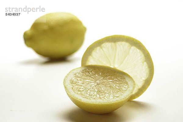 eine Zitronenhälfte und eine Zitronenscheibe mit ganzer Frucht im Hintergrund auf weißem Untergrund - Zitrone - Citrus limon - Frucht - Zitruspflanze - Zitrusfrucht - mit sehr saurem Geschmack und hohem Vitamin C-Anteil - enthält viel Pektin Phosphor Calcium Magnesium Kalium - wächst am Baum - gut für Zähne und Knochen