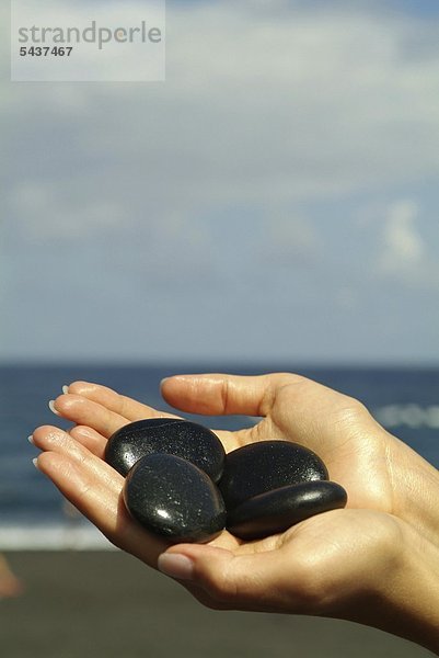 Weibliche Hände halten vier Hot Stones / Heiße Steine / La Stone - Meer in der Unschärfe im Hintergrund - La Stone - LA tibetisch für Lebensenergie   Stone = Stein - Auf 60 Grad Celsius aufgeheizte Steine   meist basaltisch   werden auf die Energiezentren des Körpers gelegt für tiefe Entspannung und neue Energie