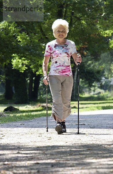 Eine ältere Frau walkt in einem Park mit Walkingstöcken auf einem Kiesweg -