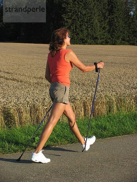 Eine Frau beim Nordic Walking geht neben einem Kornfeld auf einem asphaltierten Feldweg.