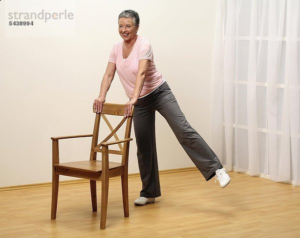 ältere Frau macht Gymnastik mit Stuhl - Bein seitlich heben - Muskelkraft - Seniorin