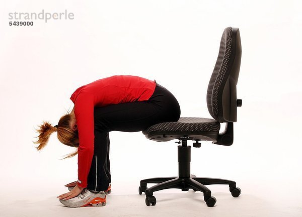 fit im Büro - junge Frau sitzt auf Bürostuhl und macht Gymnastik - Oberkörper auf Oberschenkel und Arme nach vorne herabfallen lassen - Beweglichkeit