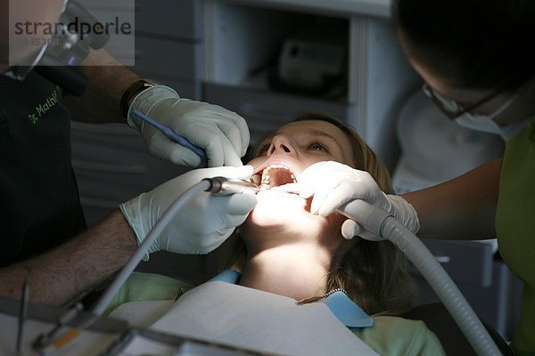 Zahnarztpraxis : Patient wird von einem Zahnarzt und einer Zahnarzthelferin mir einem Bohrer   einem Mundspiegel und Absauger behandelt