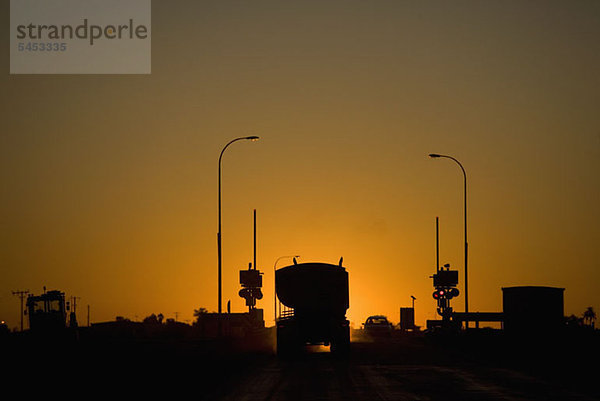 Ein Lastwagen und ein Auto halten an einem Bahnübergang  der sich gegen die untergehende Sonne abhebt.