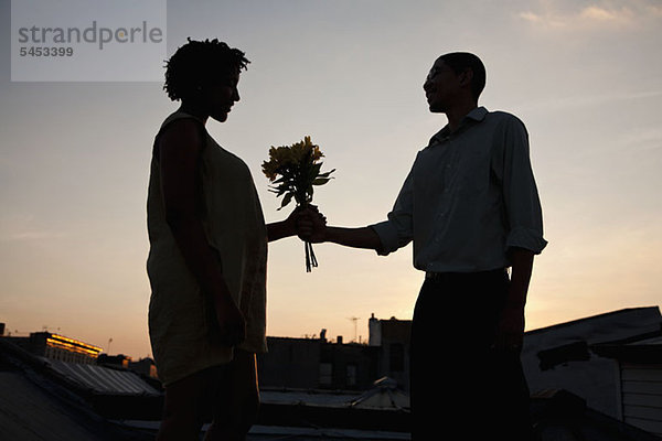 Ein Mann  der einer Frau auf einer Dachterrasse bei Sonnenuntergang Blumen schenkt.