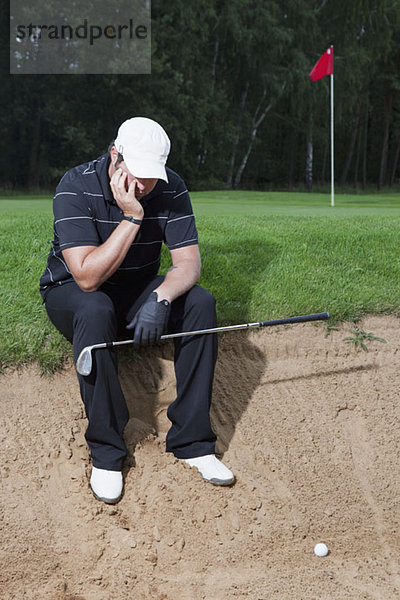 Ein unglücklicher Golfer sitzt am Rande einer Sandfalle.
