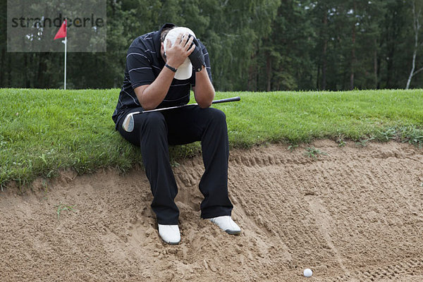 Ein Golfer sitzt am Rande einer Sandfalle  Kopf in den Händen.