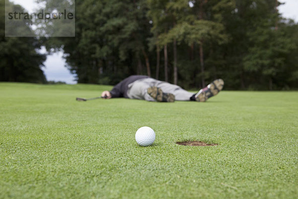 Ein verzweifelter Golfer  der auf dem Putting Green mit Ball am Lochrand liegt