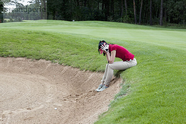 Eine Golferin sitzt am Rande einer Sandfalle  Kopf in den Händen.